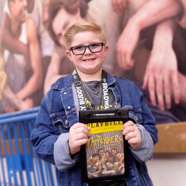 A child with glasses, proudly holding a Playbill for "The Outsiders," exudes the essence of The Broadway Buddy while wearing his denim jacket and lanyard with a badge. In the background, a photo mural decorates the wall in a gentle blur.