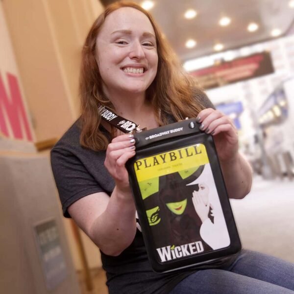 A person smiling and holding a Wicked Playbill, seated outdoors near a theater with their reliable Broadway Buddy by their side.