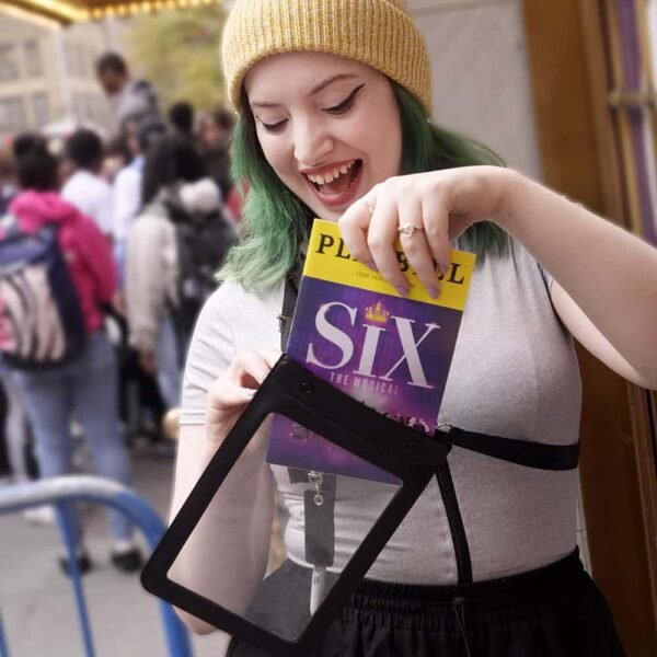 A person with green hair and a yellow beanie, known as The Broadway Buddy, proudly holds a playbill for "Six: The Musical" while standing amidst a bustling crowd.