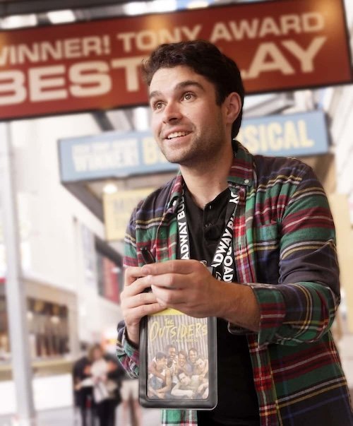 A person wearing a striped shirt holds a digital device displaying "The Outsiders." A Broadway logo lanyard is around their neck. "Tony Award Best Play" is visible in the background.