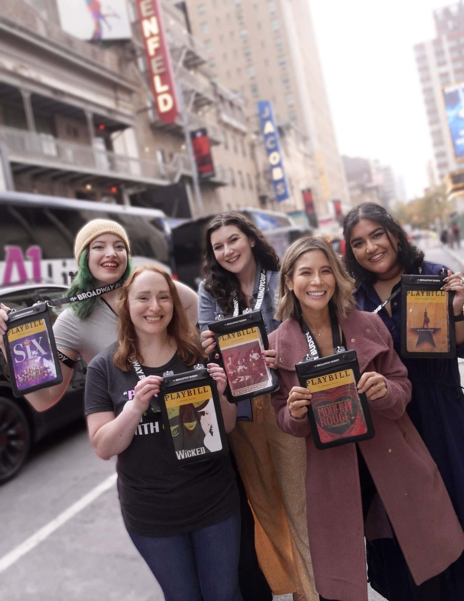 Five people stand on a city street holding theater playbills. A theater marquee and a vehicle are visible in the background.