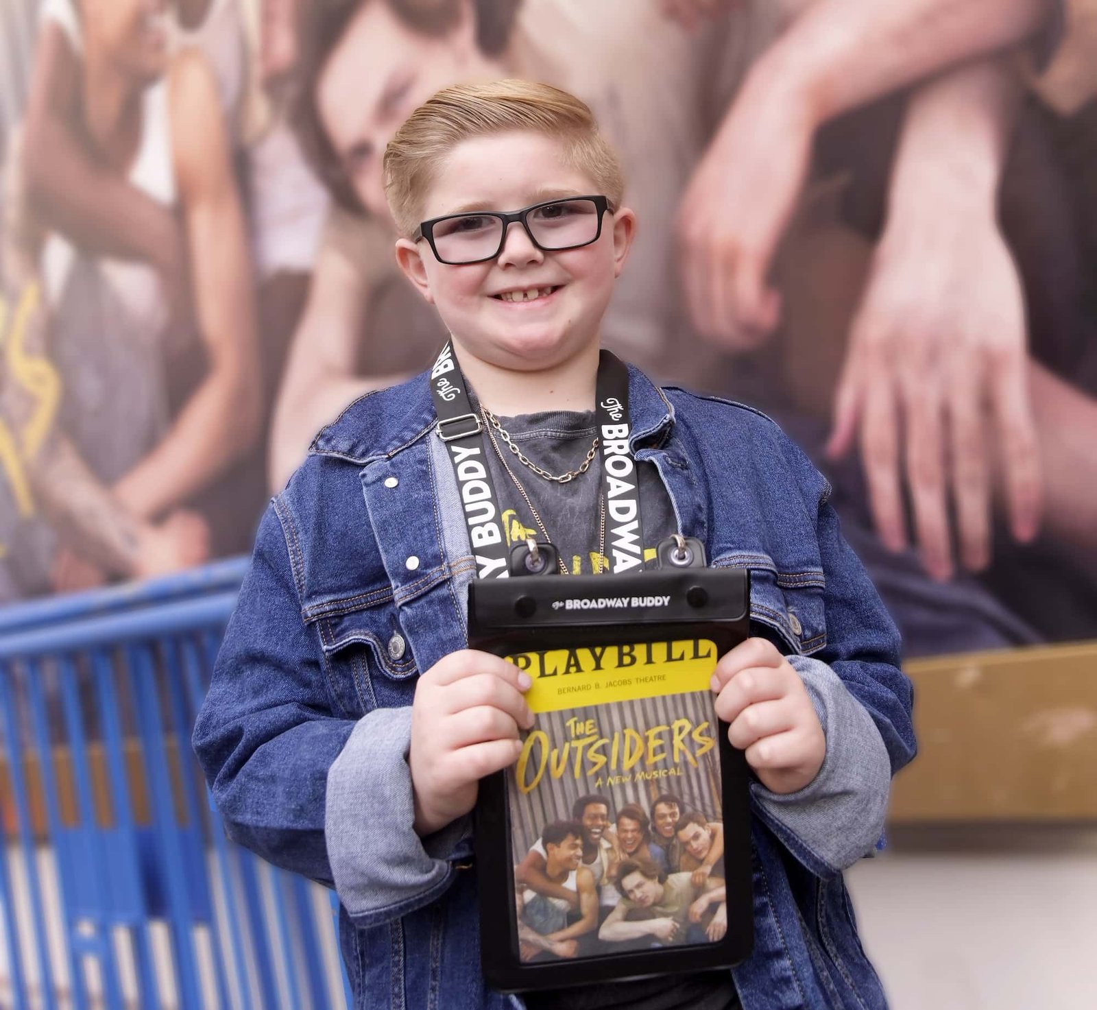 A child wearing glasses and a denim jacket holds a Playbill for "The Outsiders" in front of a blurred background.