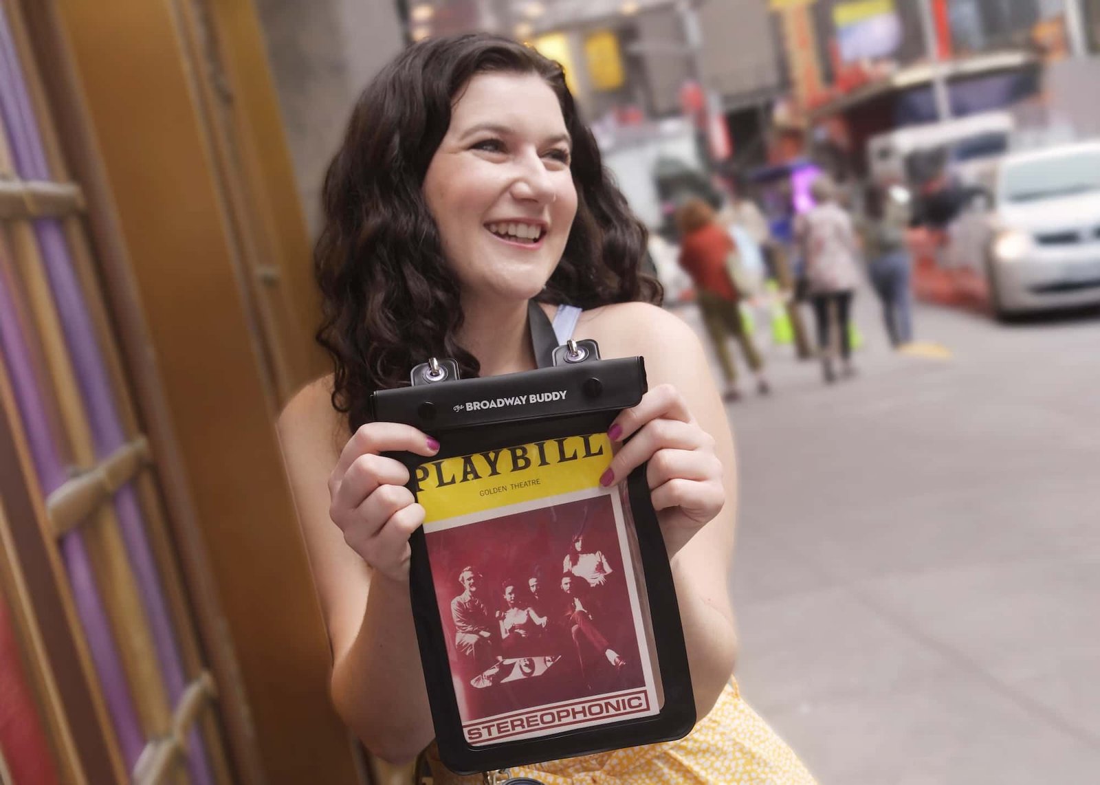 A person smiles while holding a Playbill holder labeled "Stereophonic" on a city street.