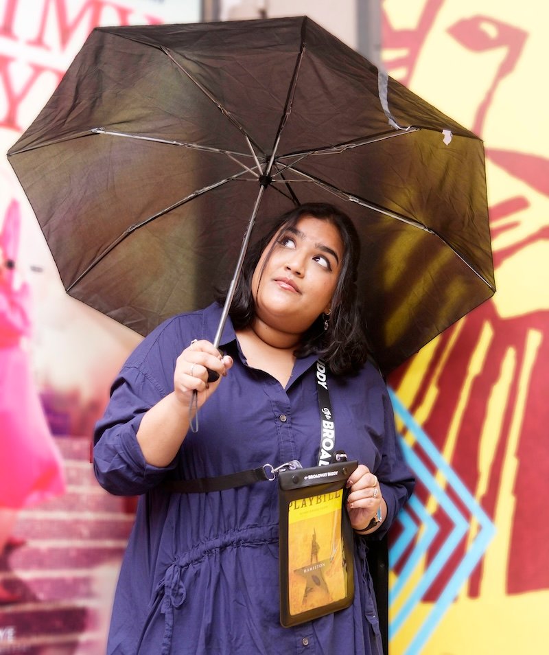 Person holding a black umbrella, wearing a dark blue coat, and looking upwards. Background features colorful geometric patterns and a blurred poster.