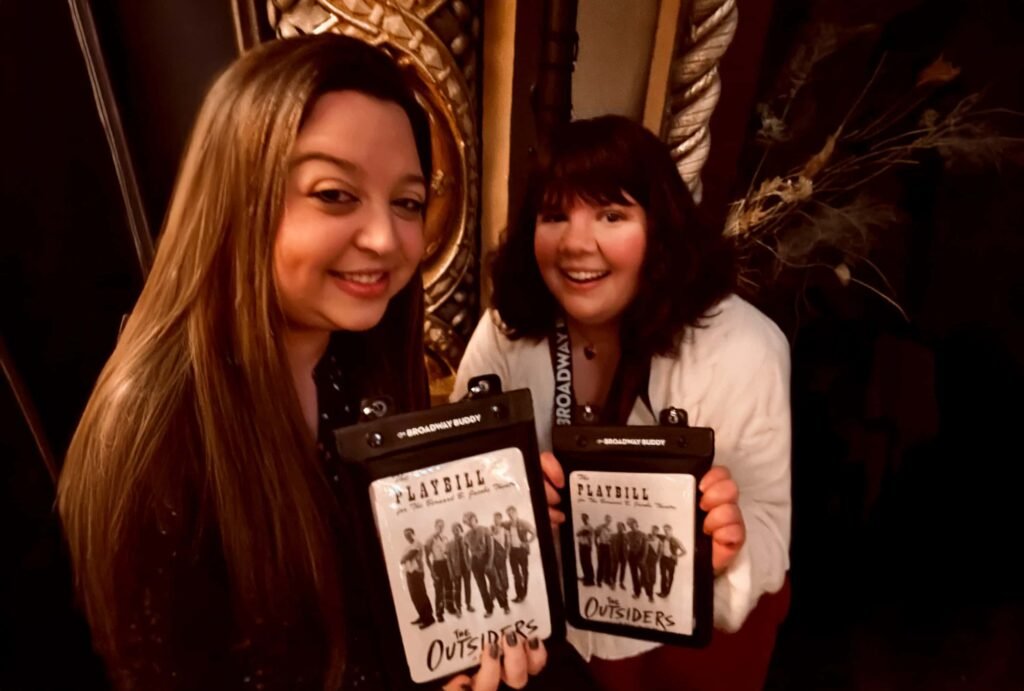 Two people smiling, each holding a Playbill for "The Outsiders." They are indoors, with decorative elements in the background.